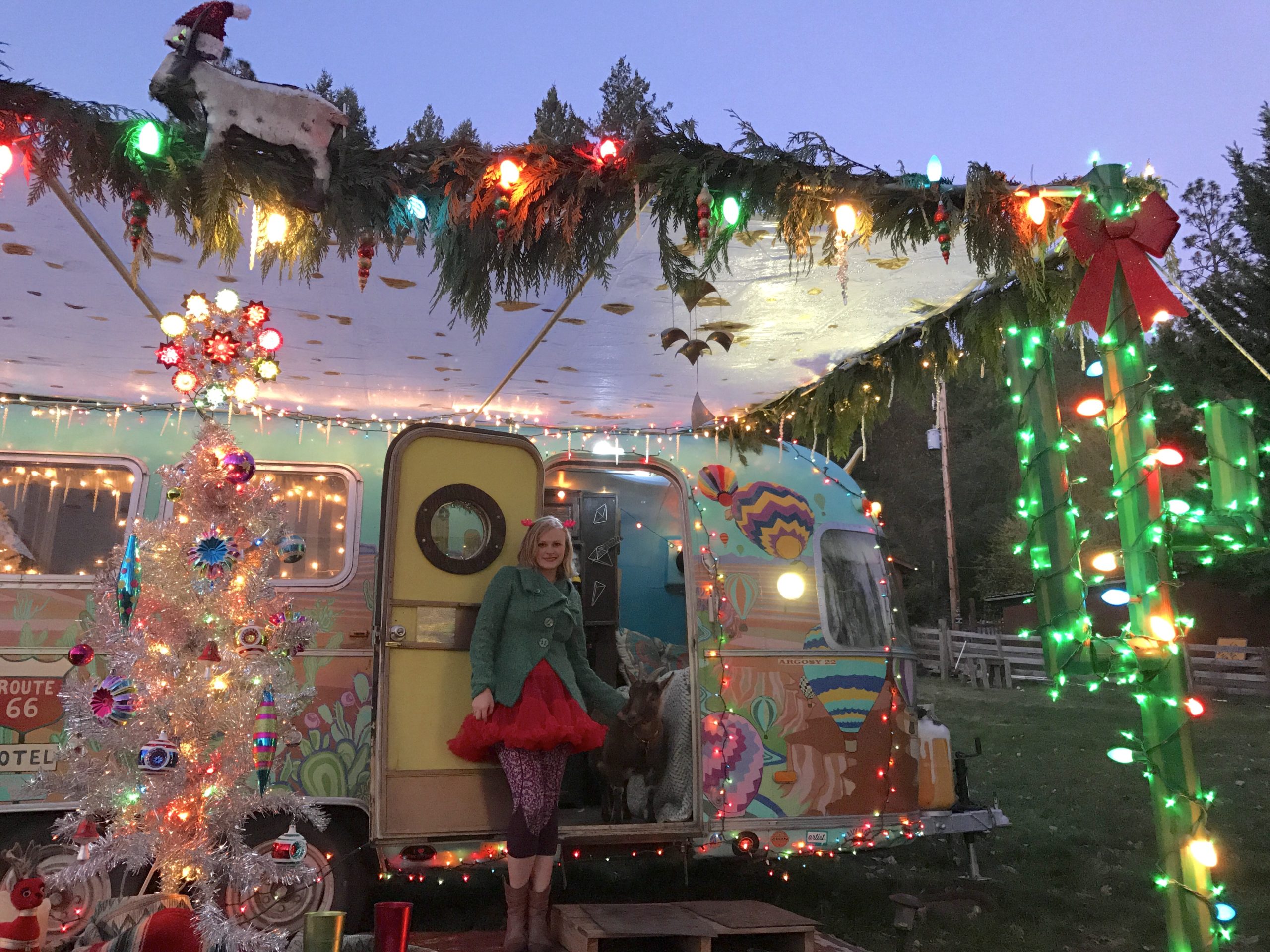 Woman with blonde hair dressed in Christmas colored skirt and coat posing in front of decorated travel trailer