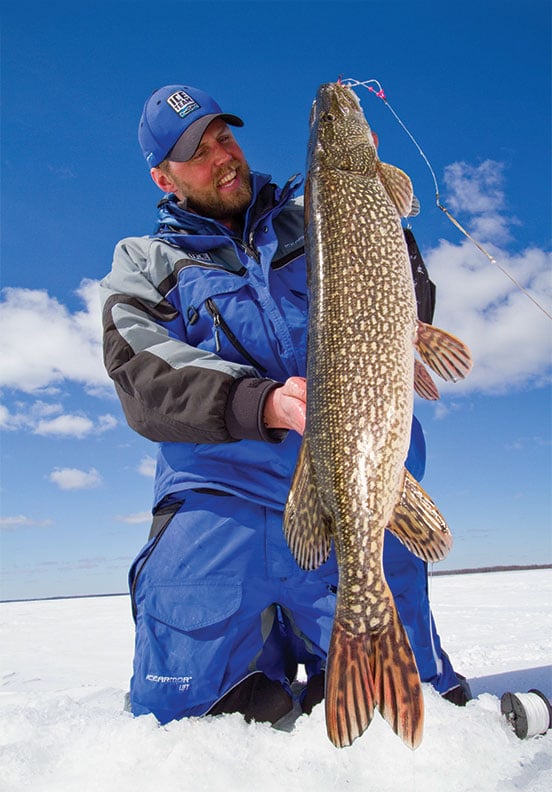 Man in blue jacket on frozen lake holding a large fish.