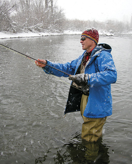 Man with hat and sunglasses in river fishing in the snow.
