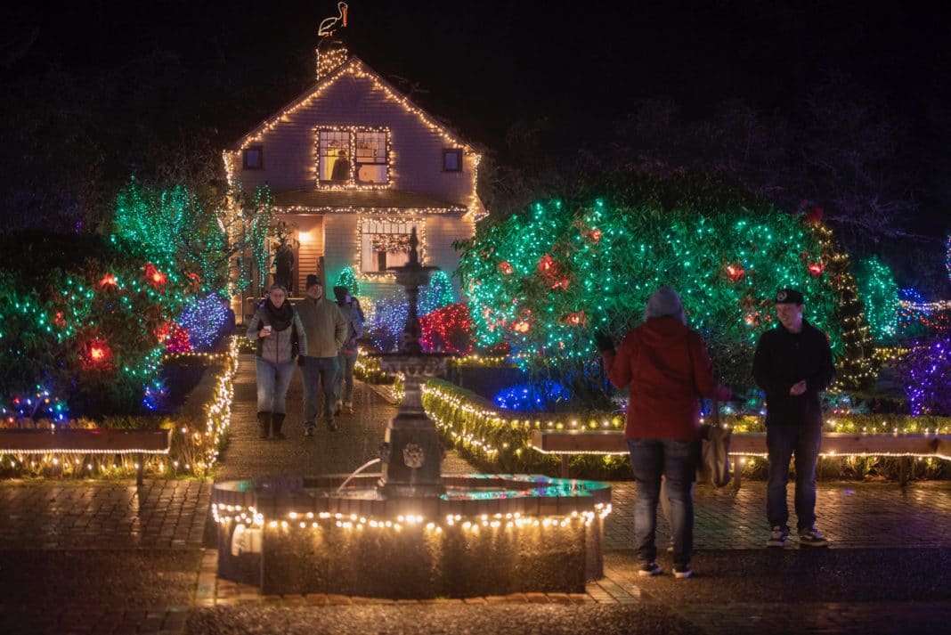 People walking along street near houses covered in holiday lights, dressed warmly 