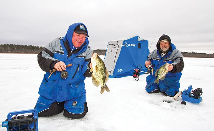 Man in blue parka on the ice with a fish in his hand and another man and tent in the background