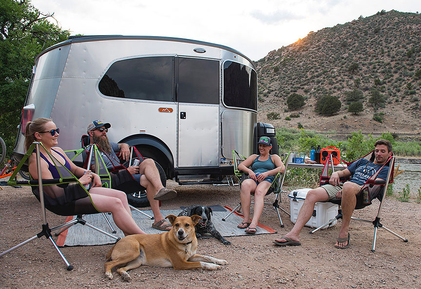 Four people in camp chairs and a dog in front of the Basecamp trailer.