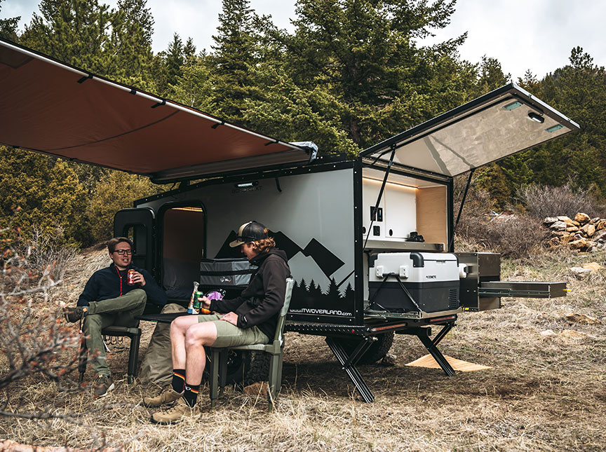 Two people in camp chairs in front of trailer with awning extended.