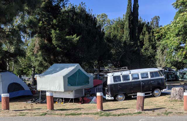 A black-and-white 1966 VW bus and 1969 Heilite pop-up camper under the shade of trees.