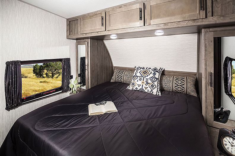 Trailer bedroom with dark bedspread on queen bed and cabinets above