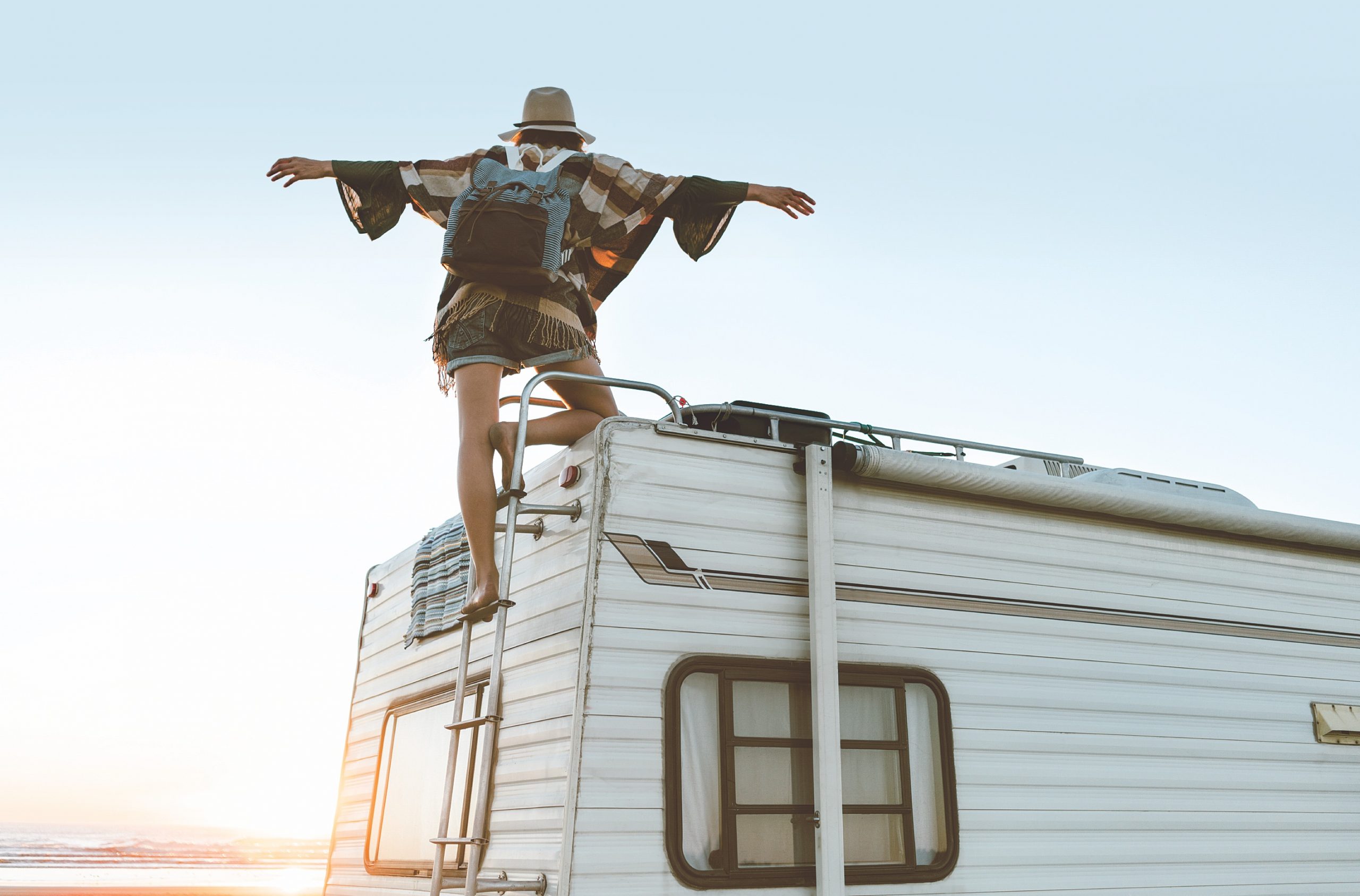 Charming girl with hat, poncho, backpack standing on roof of recreational vehicle on the ocean beach at sunset.