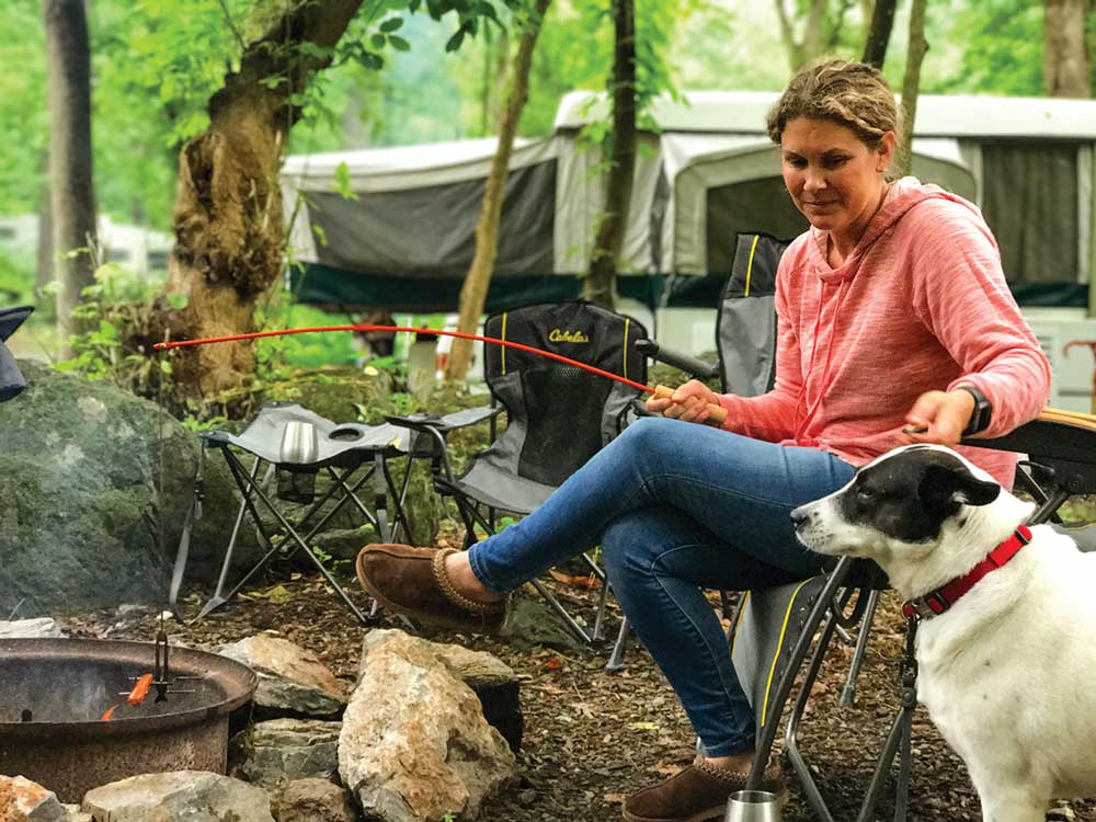 Woman holding stick over campfire looking at dog with trailer in background