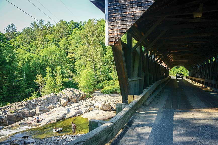 In the town of Bath, a car crosses the 158-foot-long Swiftwater Bridge, built in 1849, as bathers sun themselves on the rocks of the Wild Ammonoosuc River below the historic span.