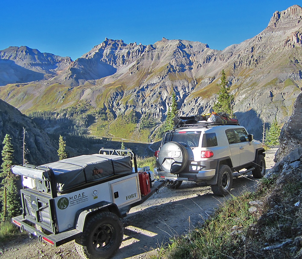 Silver SUV with camping gear attached on top towing 2-wheel MOAB travel trailer in mountains