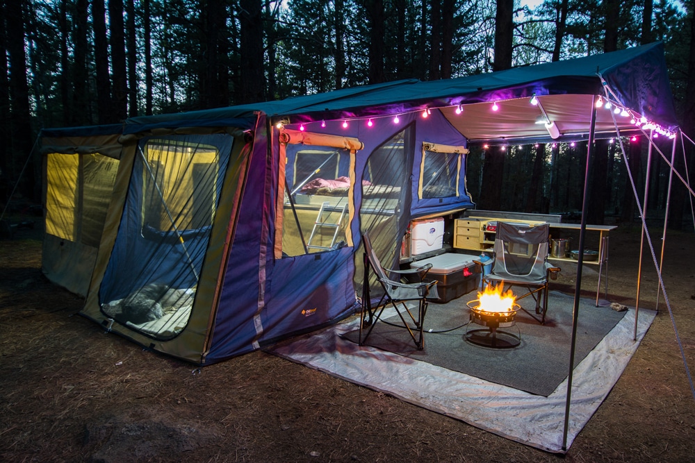 Colorful tent with awning lit up with purple lights and fire pit in woods