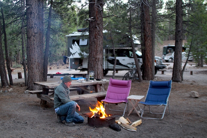 Campfire, two chairs and the author's husband at a wooded campsite.