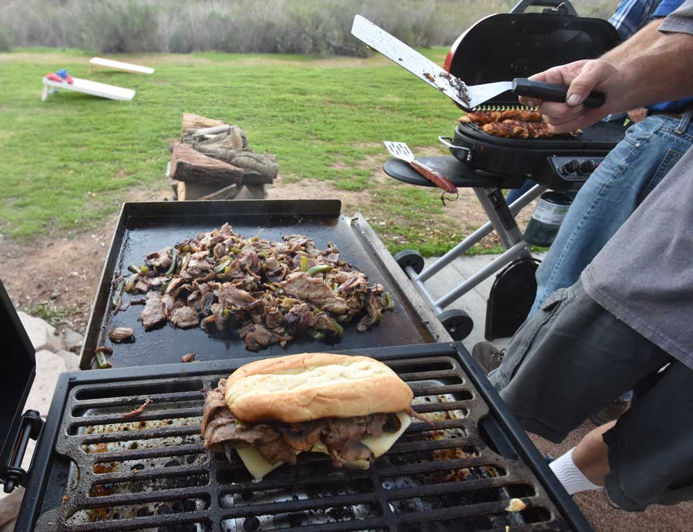 A philly cheese steak sandwich warms on the blackstone tailgater grill while more ingredients are cooked on the griddle side of the grill