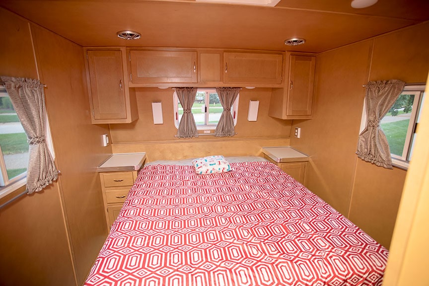 Walk-around queen bed with red and white bedspread and paneled walls in Holiday House trailer