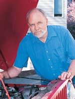 Man with grey beard and wearing blue shirt leaning over open car hood