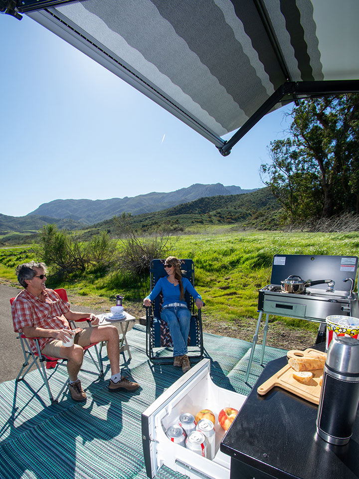 Two RVers relaxing under rear awning of Lance 2075 travel trailer with outdoor kitchen