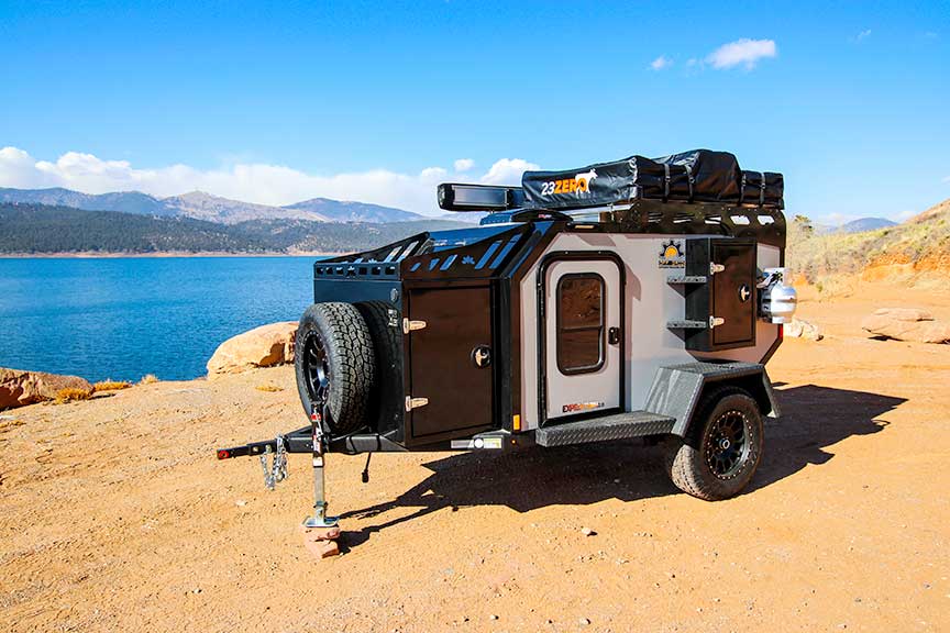 Expedition trailer on sand in front of blue water.