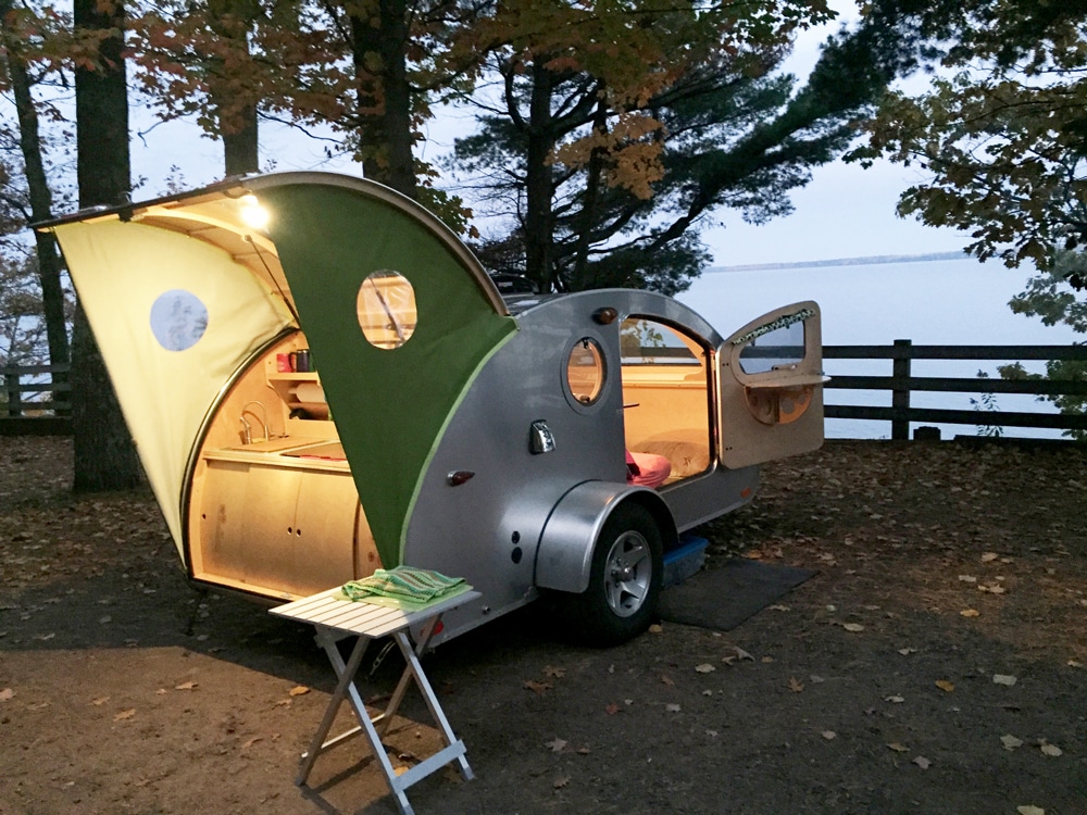 Green and silver teardrop camper parked along water with trees