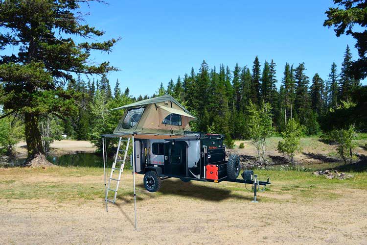 Trail Head trailer with rooftop tent up and evergreen trees behind it.