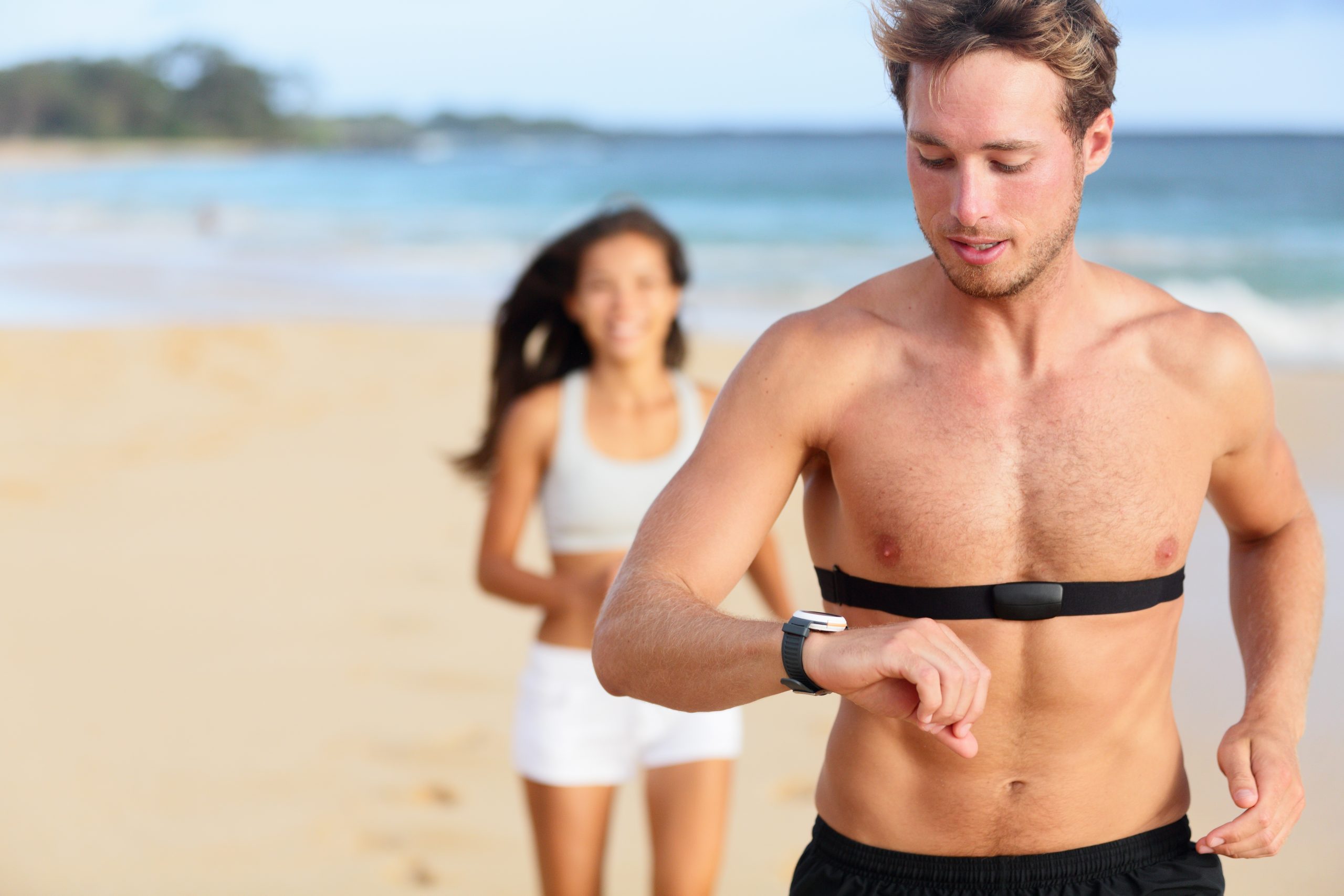 Running young man jogging on the beach using a heart rate monitor.