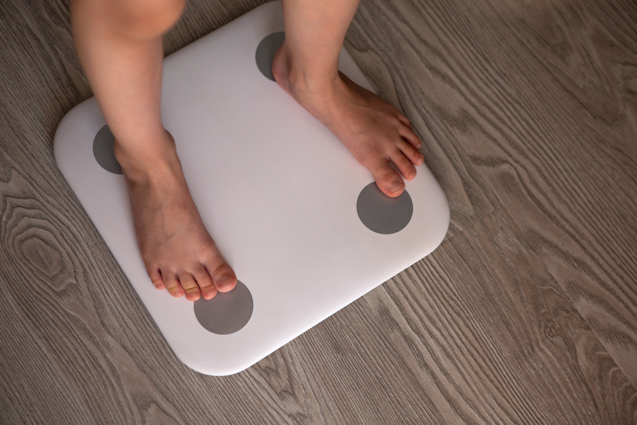 Kid girl or boy stands and weighs himself on white modern electronic smart scale. Scales stand on grey wooden floor. Only legs are visible. Concept of Internet of things, weight control, health.