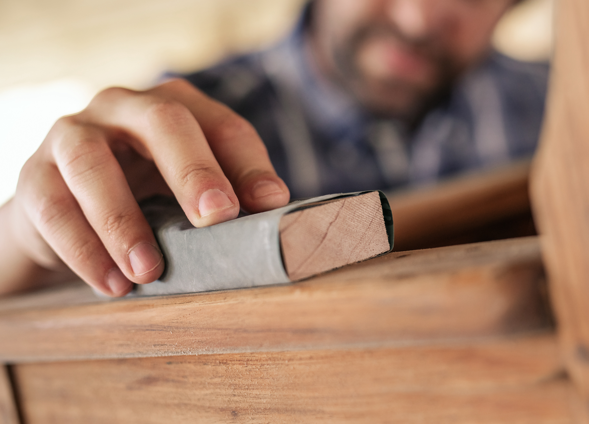 Closeup of hand of woodworker using sandpaper to sand a wooden chair while working in his woodworking shop