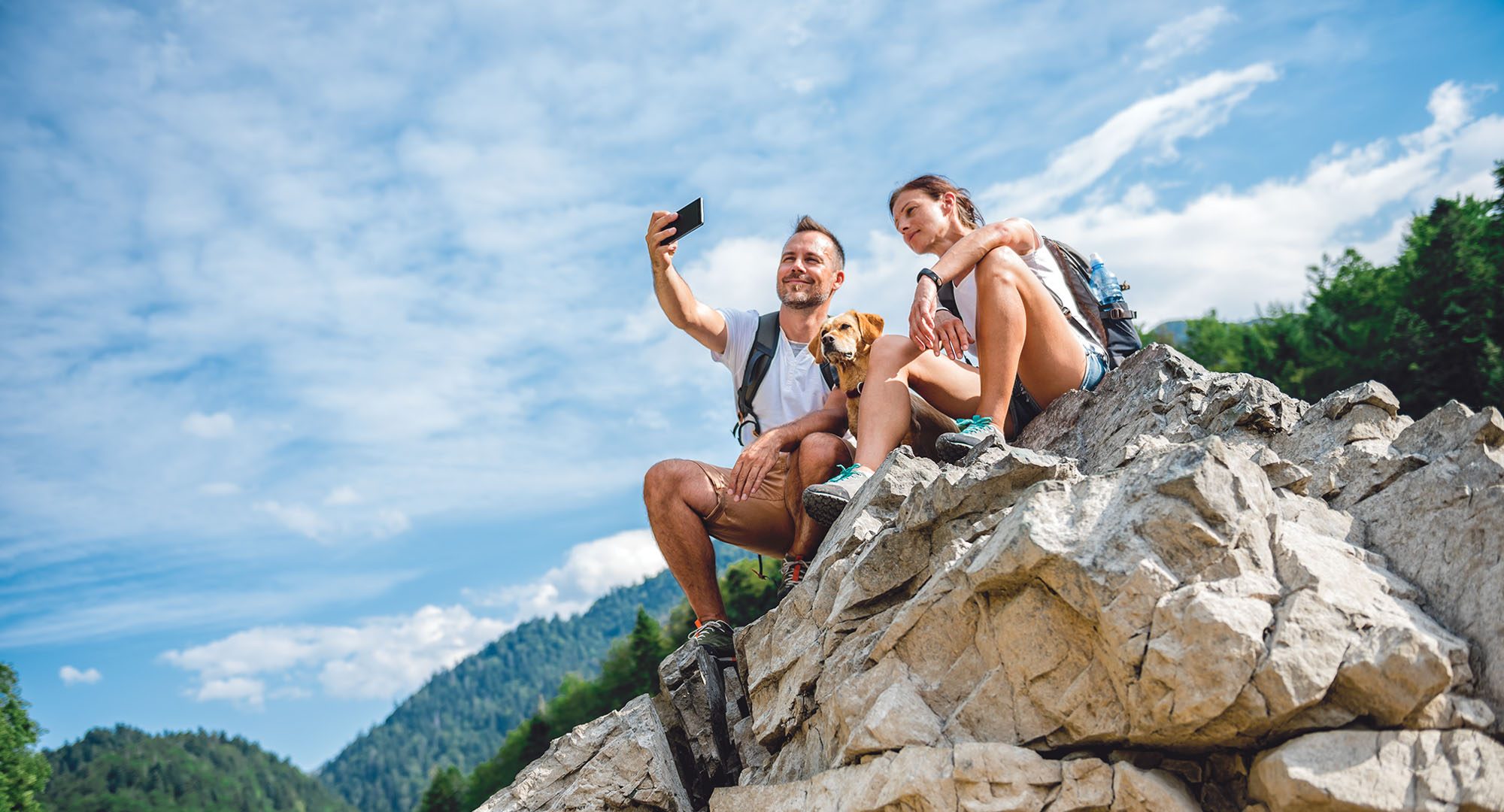 Hiker couple with small yellow dog sitting on mountain peak and taking picture with smart phone