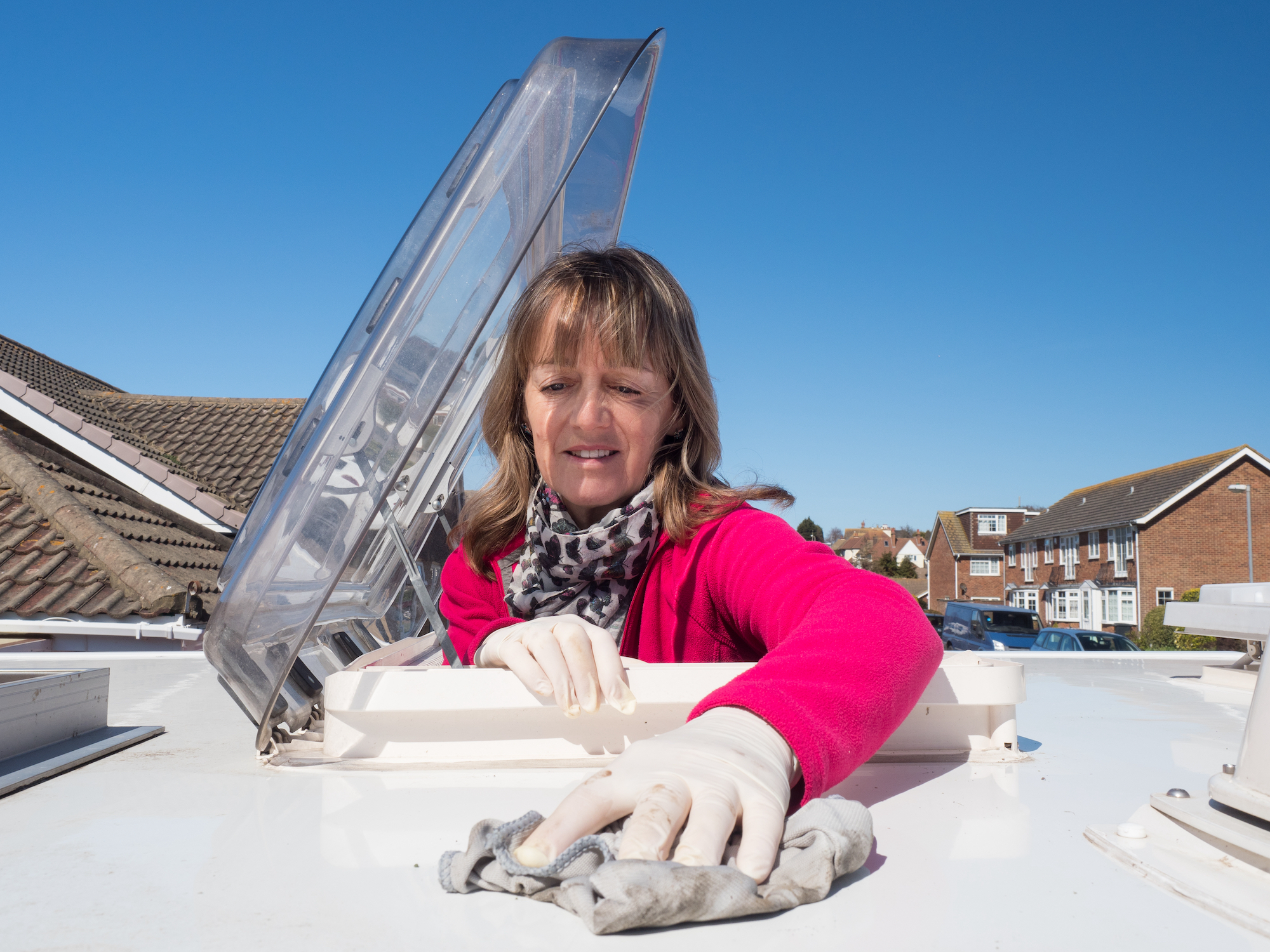 A lady motorhome owner cleans the roof of her vehicle with latex gloves on.The top half of her body is visible as she has climbed through the roof skylight.Vivid pink fleece.Amusing.Image