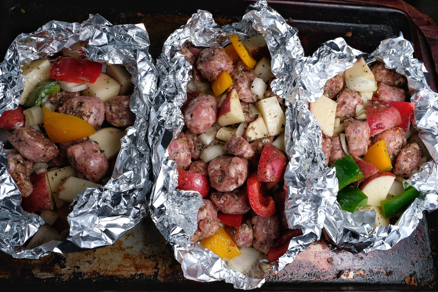 Three foil packets with cut sausages, vegetables, oil and pieces ready to put in oven or grill