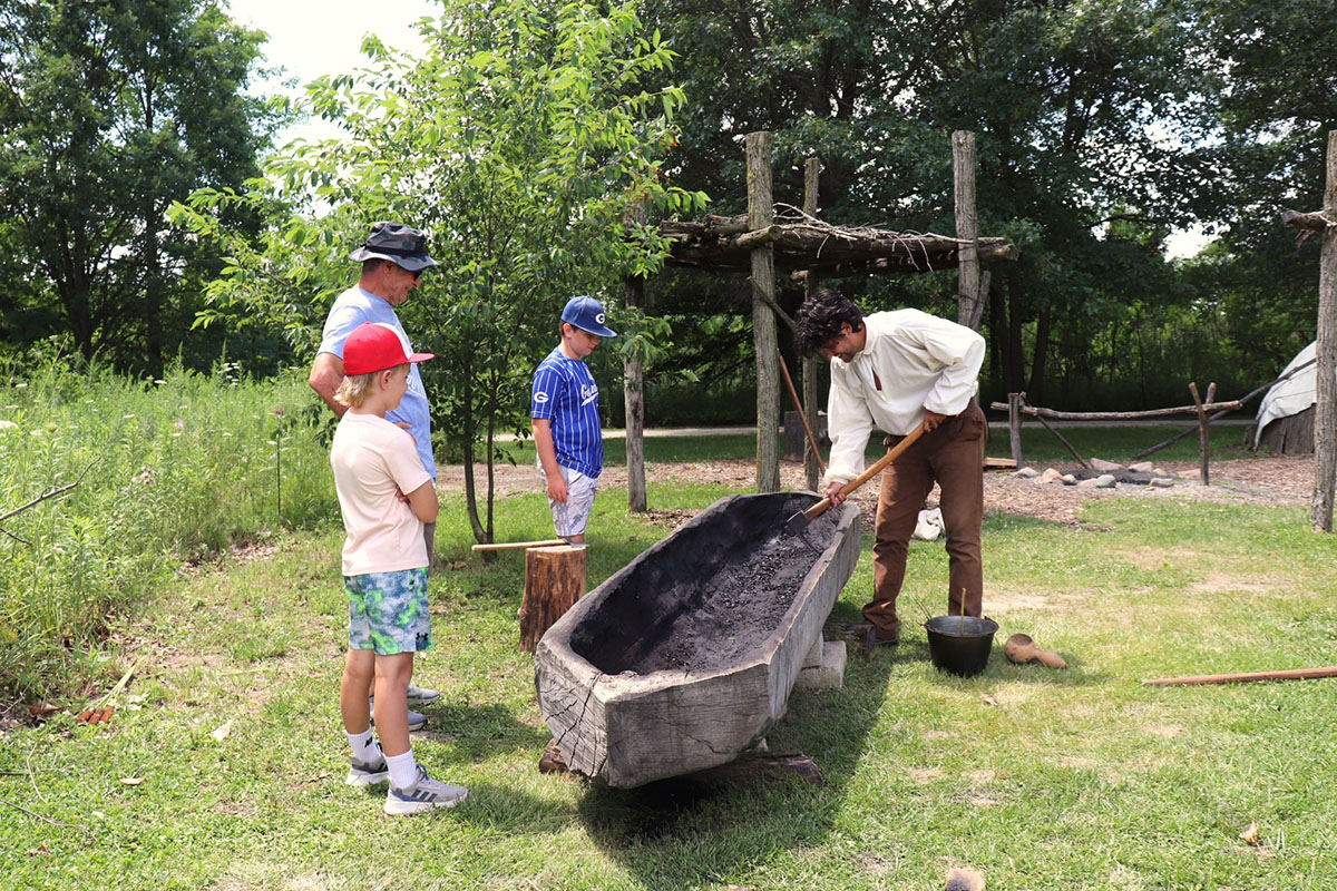Man dressed in 1800s period costume make dugout canoe.