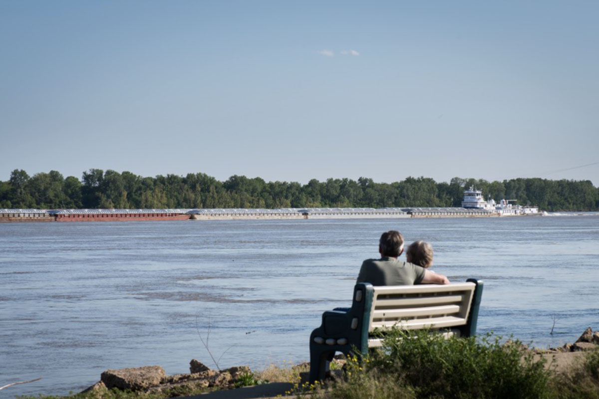 A couple on a park bench watch a long barge.